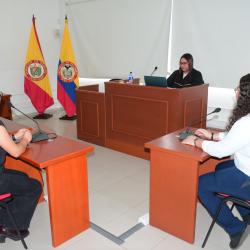 Fotografia muestra a tres personas en lo que parece ser una audiencia judicial o una reunión formal. Una mujer con una toga negra está sentada en el centro, detrás de un escritorio elevado con dos banderas colombianas. Dos mujeres están sentadas a los lados, frente a ella. Todos tienen micrófonos en sus escritorios.