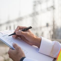 Imagen que muestra unas Manos de un trabajador con uniforme blanco y chaleco reflectante escribiendo en un portapapeles con una pluma negra