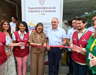 Un grupo de personas sonrientes cortando una cinta roja frente a un cartel de la "Superintendencia de Industria y Comercio". A la izquierda, un ícono de ubicación con una casa.
